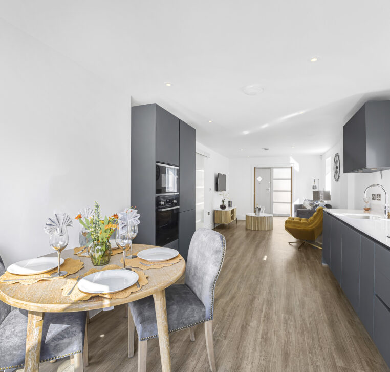 Long view of a contract kitchen with grey units and built-in appliances, opening to a dining space with outdoor access