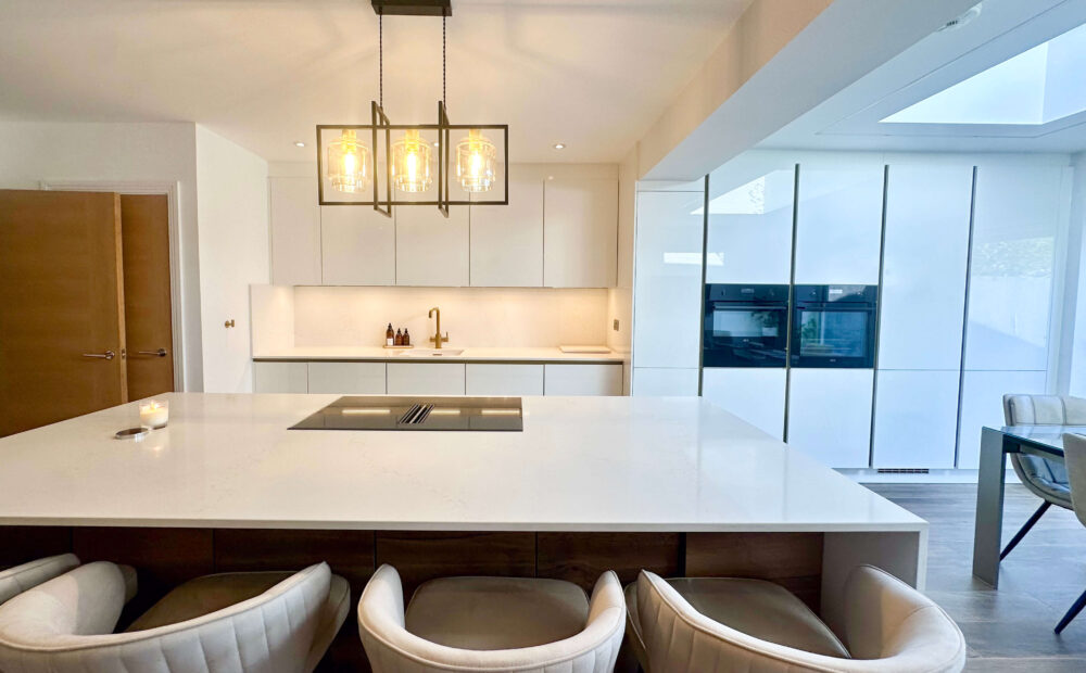 Sleek white kitchen with a large waterfall island, pendant lighting, a gold faucet, and built-in ovens reflecting a contemporary aesthetic