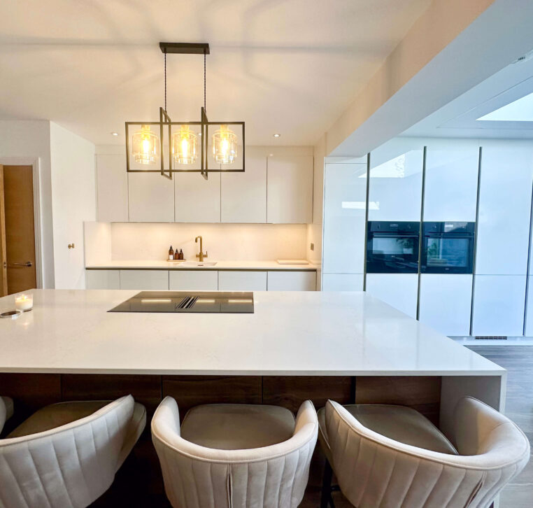 Sleek white kitchen with a large waterfall island, pendant lighting, a gold faucet, and built-in ovens reflecting a contemporary aesthetic