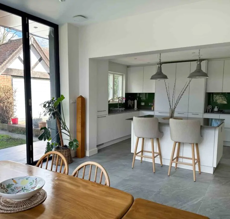 A minimalist kitchen featuring a white island, light grey bar stools, industrial pendant lighting, and a dark countertop.