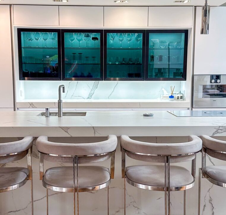 A contemporary kitchen with a glass-fronted cabinet, white marble worktops, and soft ambient lighting.