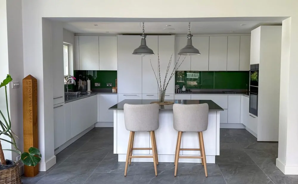 A modern white kitchen with a sleek island, green glass backsplash, and two upholstered bar stools