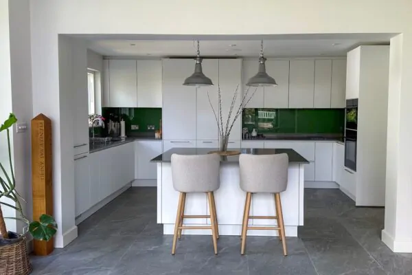 A modern white kitchen with a sleek island, green glass backsplash, and two upholstered bar stools