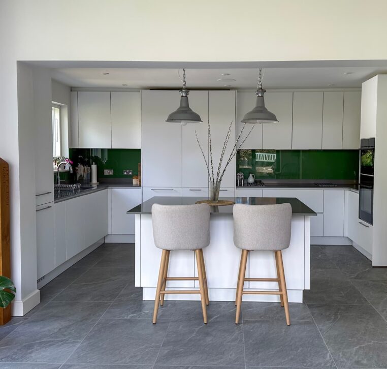 A modern white kitchen with a sleek island, green glass backsplash, and two upholstered bar stools