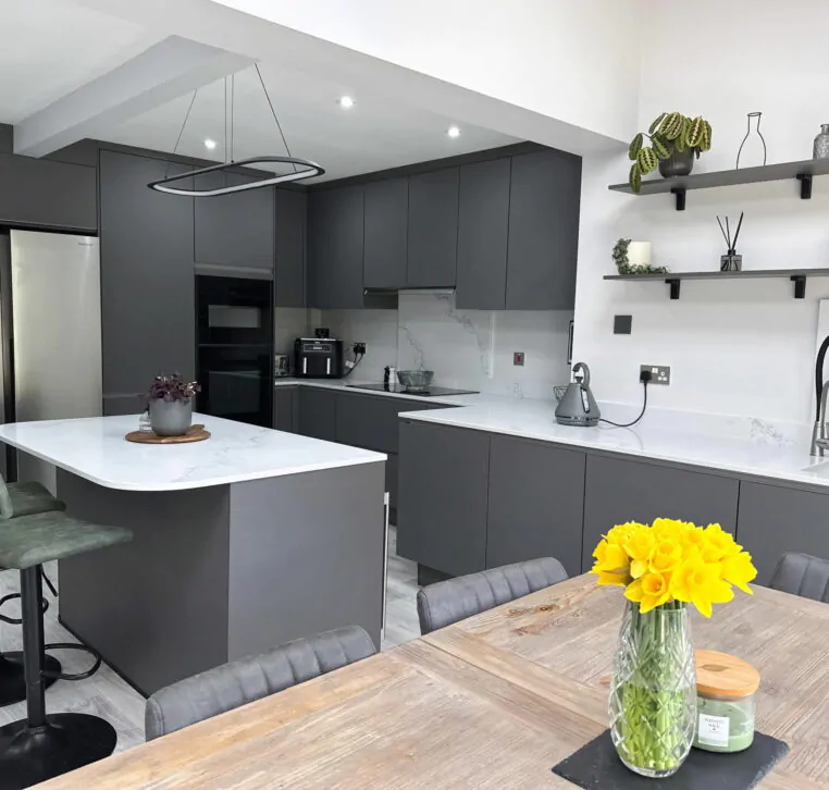 Modern grey kitchen with a white quartz island, open shelving, and dining area with large garden doors.