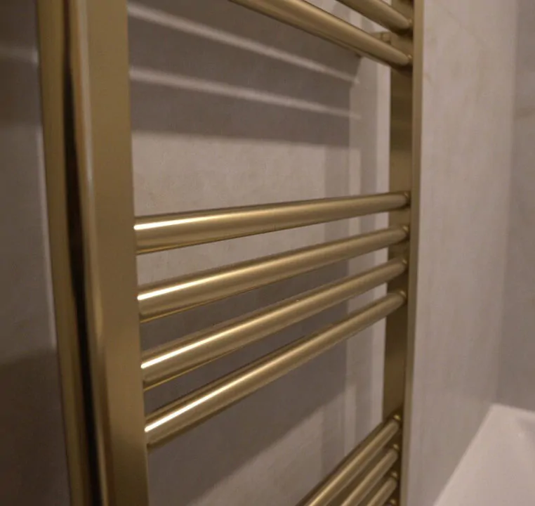 Close up of a designer brushed gold heated towel rail installed on a marble effect tiled bathroom wall.