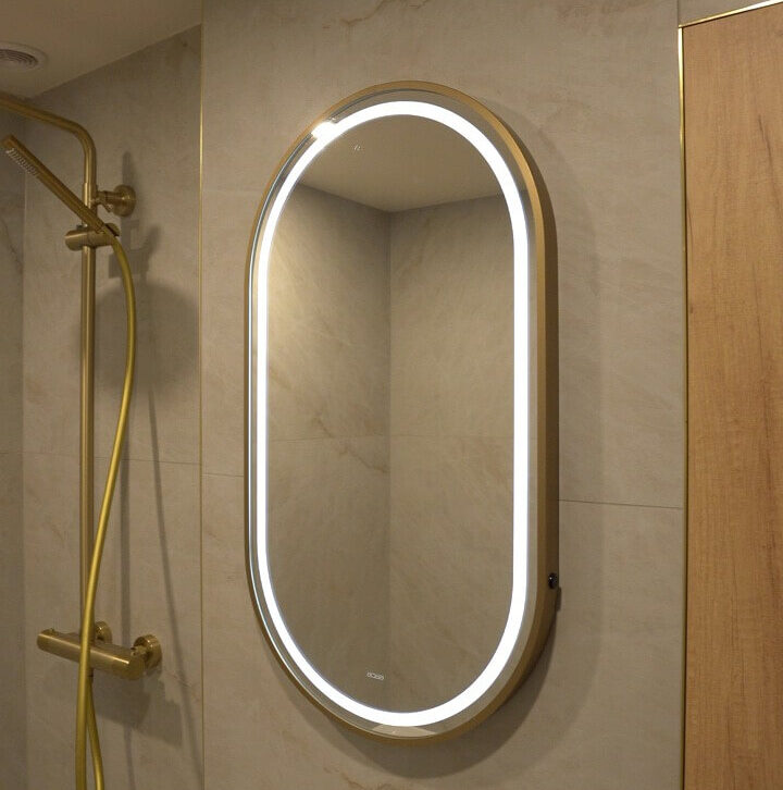 Oval LED backlit bathroom mirror with a brushed gold frame mounted on a neutral stone-effect tiled wall.