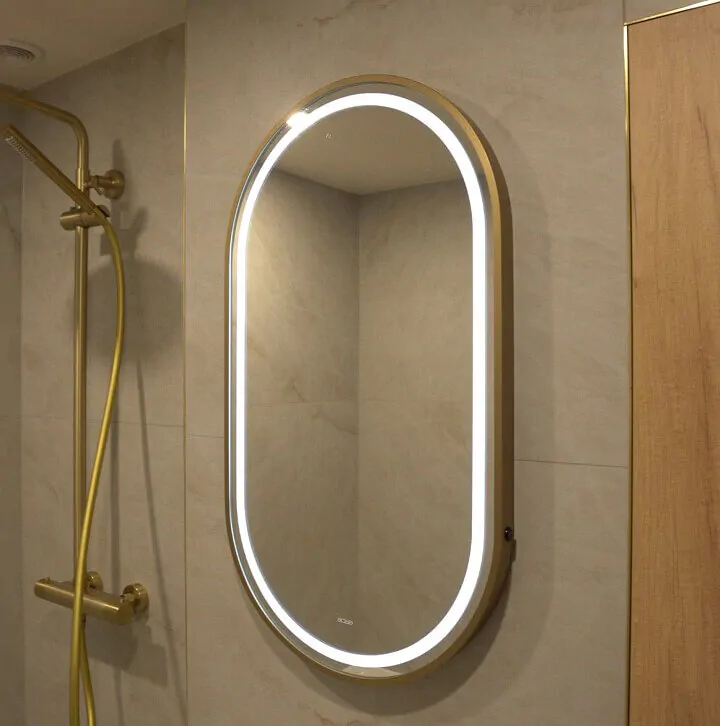 Oval LED backlit bathroom mirror with a brushed gold frame mounted on a neutral stone-effect tiled wall.