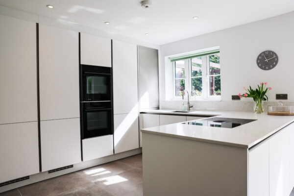 White handleless kitchen with large island-style worktop, integrated hob, built-in ovens and garden views