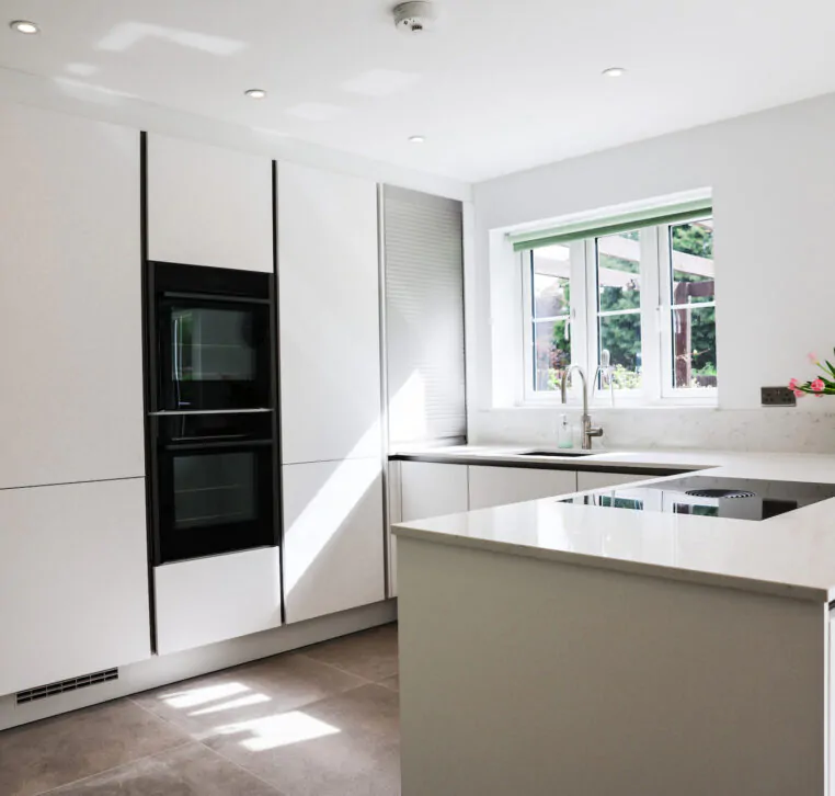 White handleless kitchen with large island-style worktop, integrated hob, built-in ovens and garden views