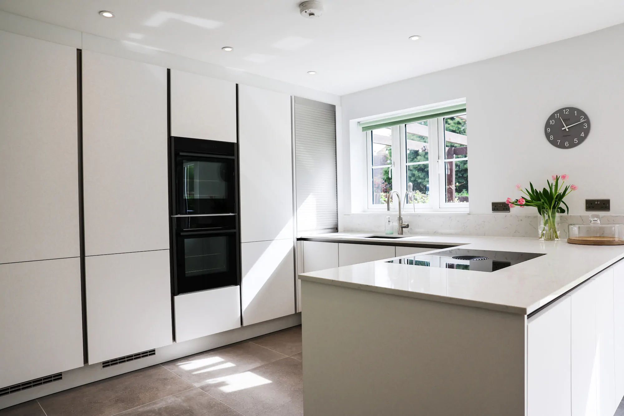 White handleless kitchen with large island-style worktop, integrated hob, built-in ovens and garden views